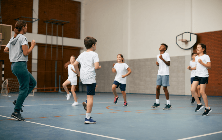 Kinder führen gemeinsam mit einer Sportlehrerin Lauf- und Sprungbewegungen in einer Turnhalle aus.