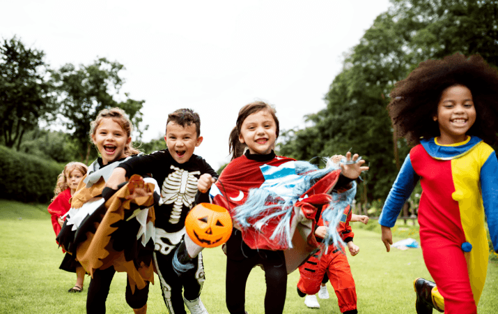 Eine Gruppe lachender Kinder in bunten Halloween-Kostümen – darunter ein Skelett, ein Clown und ein Umhang-Träger mit Kürbiseimer – rennt fröhlich über eine grüne Wiese in einem Park.