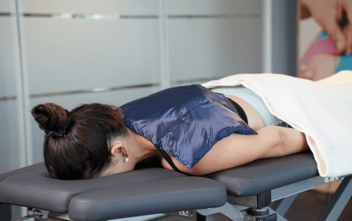 Frau liegt auf einer Physiotherapie-Behandlungsliege mit einer dunkelblauen Wärmepackung auf Nacken und oberem Rücken.