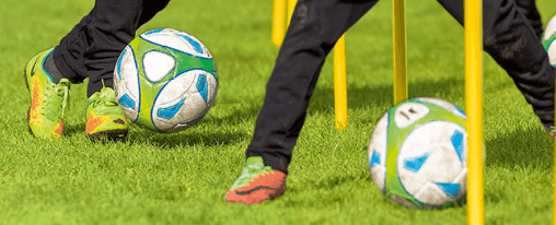 Fußball-Ausrüstung kaufen Zwei Fußballspieler beim Dribbeltraining mit Fußbällen um Stangen auf einem Sportplatz