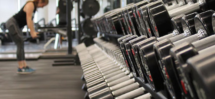 Ein Regal mit Hanteln in einem Fitnessstudio, im Hintergrund führt eine Athletin Übungen mit Hanteln aus