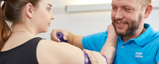 Therapeut in einem blauen Poloshirt behandelt Patientin mit lila Faszienrolle am Oberarm