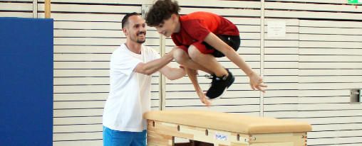Kind springt über einen Kübler Sport Sprungkasten mit Hilfestellung eines Sportlehrers