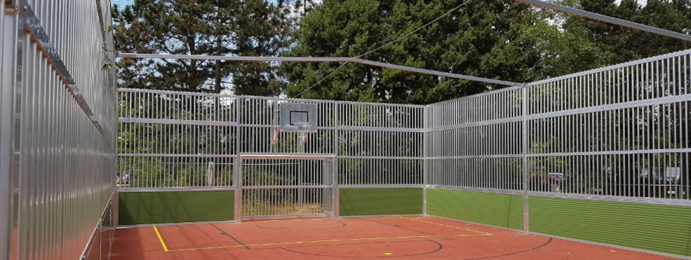 Cage Soccer Court Arena aus Metall, grünen Banden und orangenen Boden und einem Minitor sowie Basketballbrett