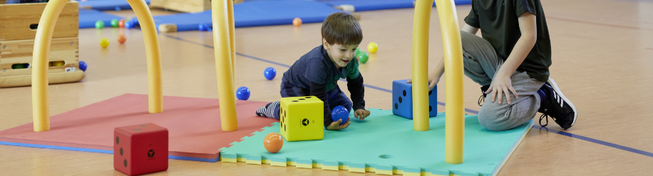 Kinder spielen auf einer Spielmatte mit Spielbällen und Schaumstoffwürfel in einer Turnhalle
